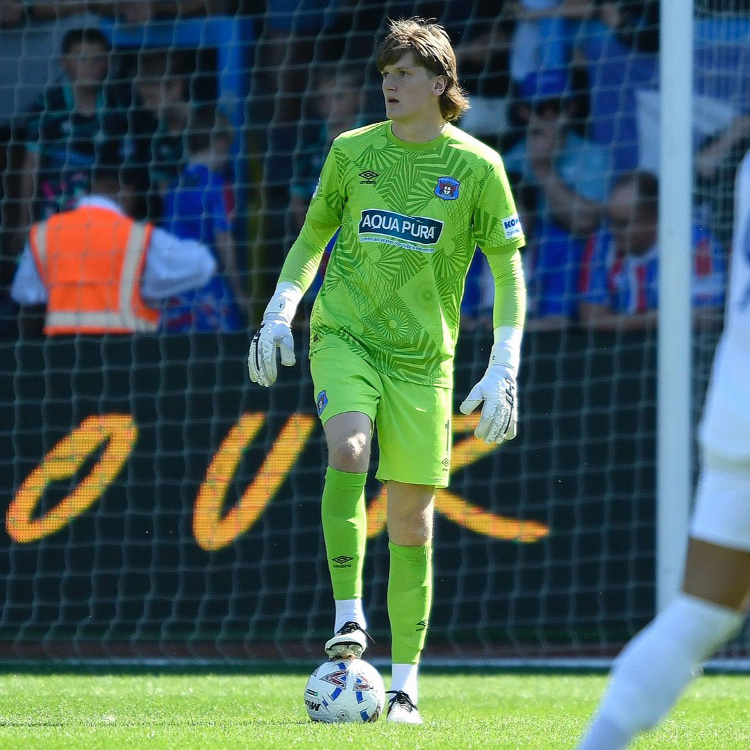 Goalkeeper in green kit standing on a soccer field with fans and advertising boards in the background.